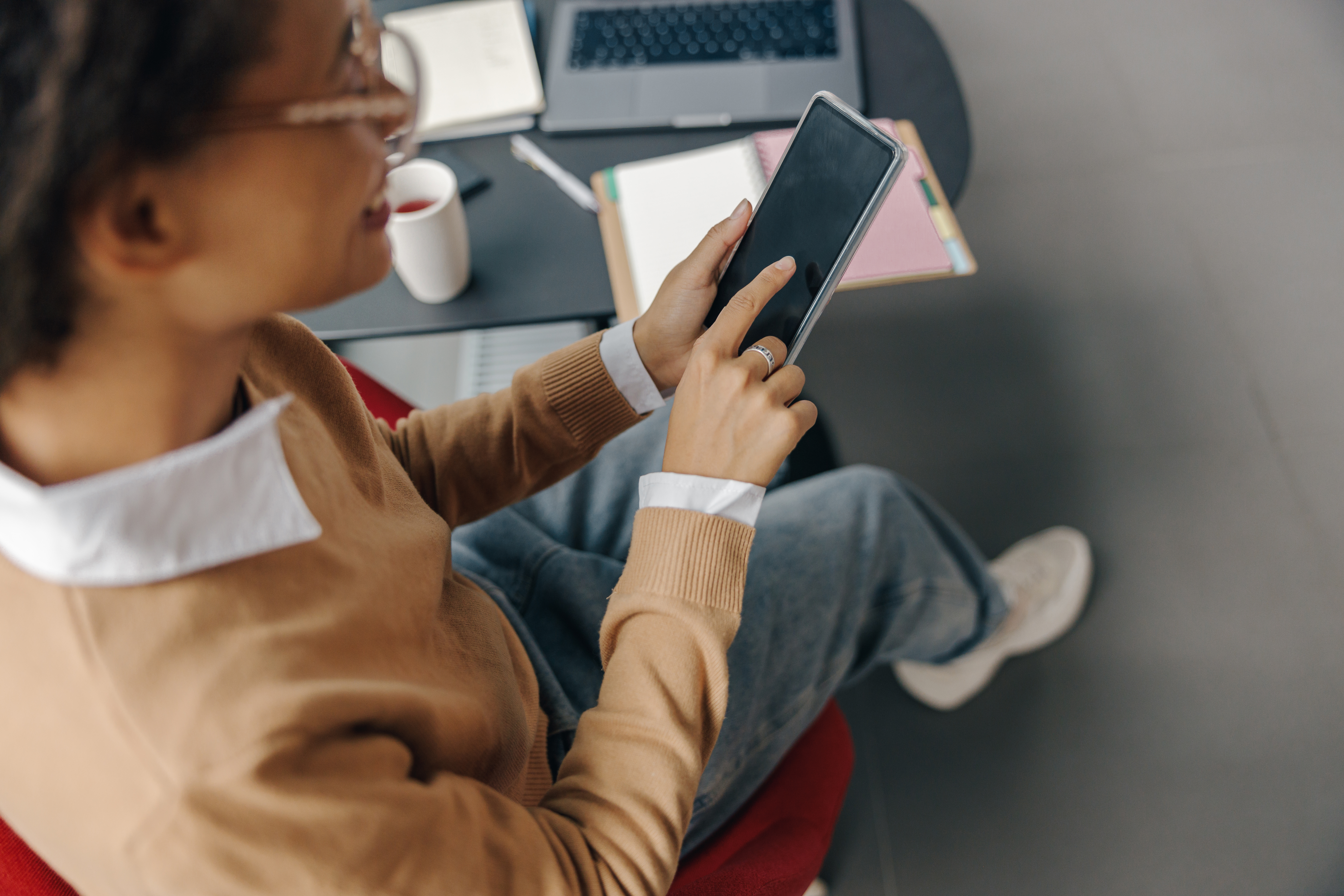 Person sitting in a cozy workspace using a smartphone for mobile learning, with a laptop, notebook, and coffee nearby, suggesting a flexible and modern working environment.