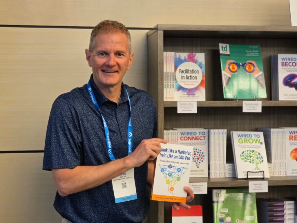Mike Taylor smiles while holding his co-authored instructional design book, Think Like a Marketer, Train Like an L&D Pro, in front of a display of learning and development books.