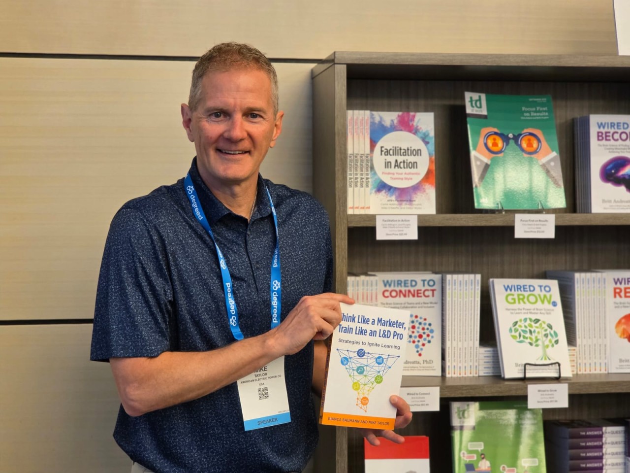 Mike Taylor smiles while holding his co-authored instructional design book, Think Like a Marketer, Train Like an L&D Pro, in front of a display of learning and development books.
